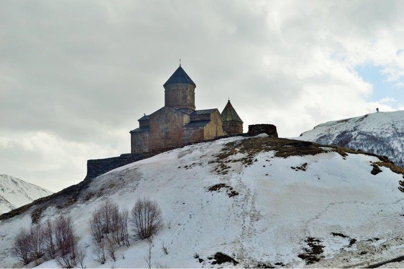A small domed church on a snow-covered hilltop with the high Caucasus rising behind