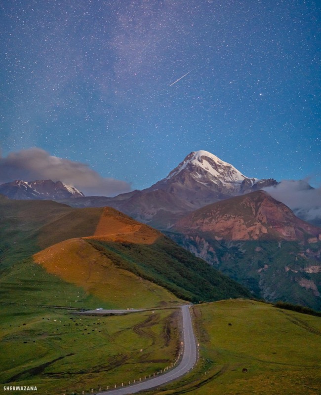 A snow-dusted Caucasus peak rising above green foothills and a winding mountain road