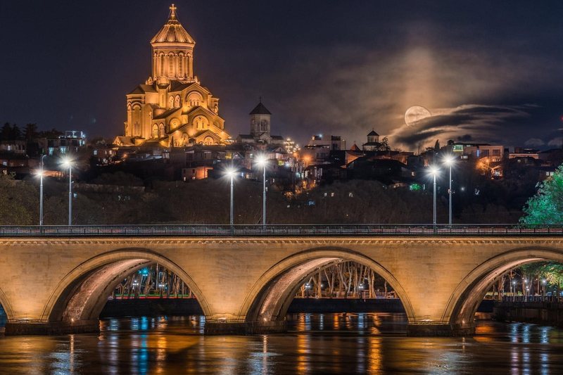 A floodlit cathedral and an illuminated bridge over the Mtkvari river in Tbilisi at night